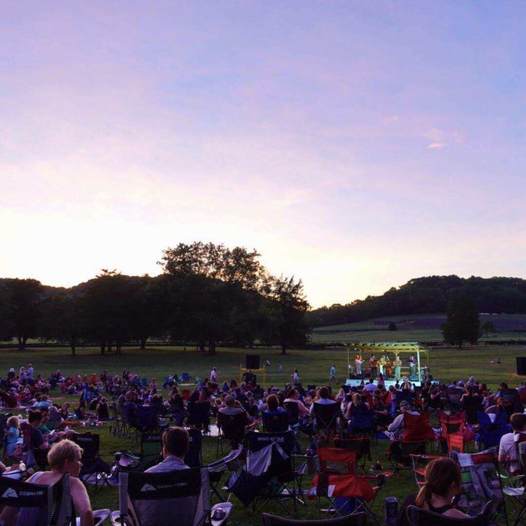 People in folding chairs in a park at dusk, attending an outdoor concert with a stage in the background under a colorful sky.