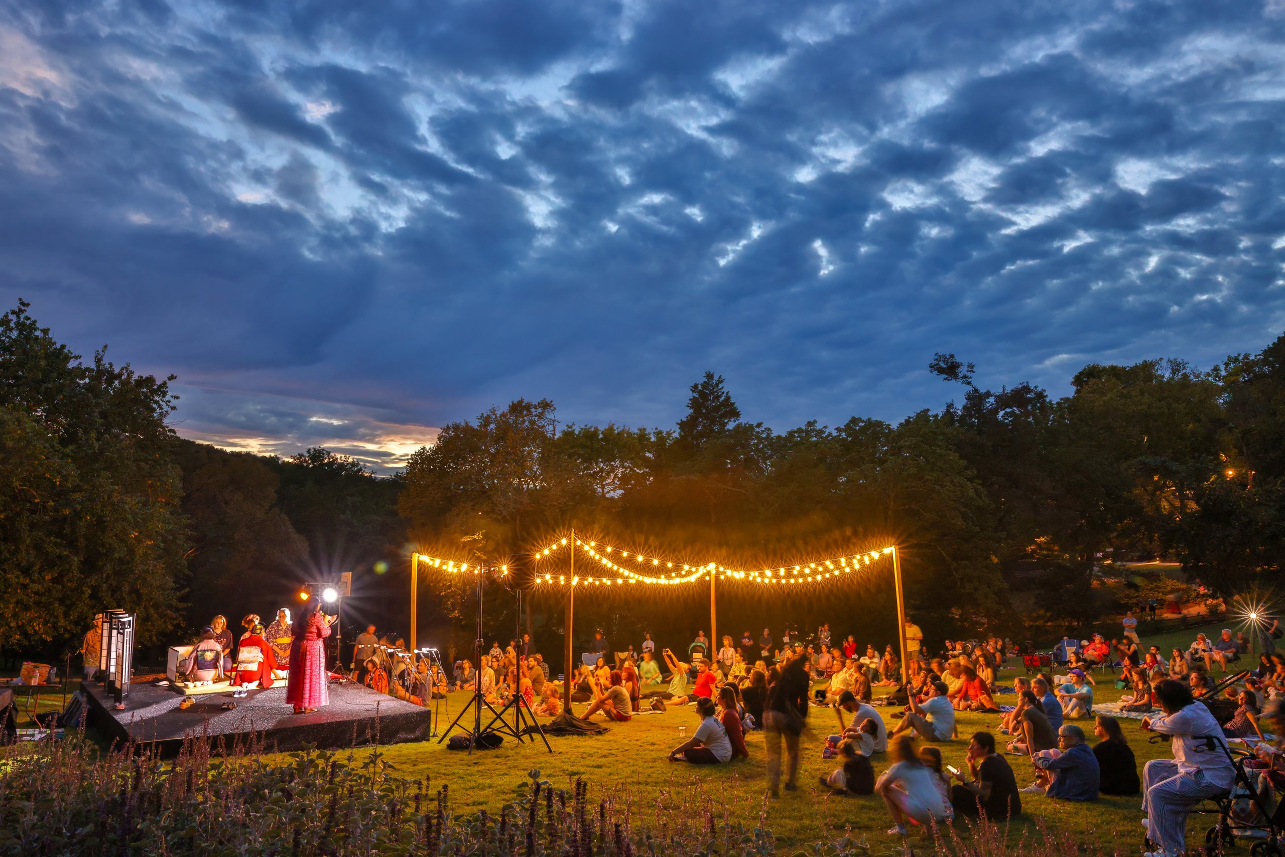 people sitting in lawn at Cheekwood garden beneath canopy of lights watching stage with Japanese musicians.