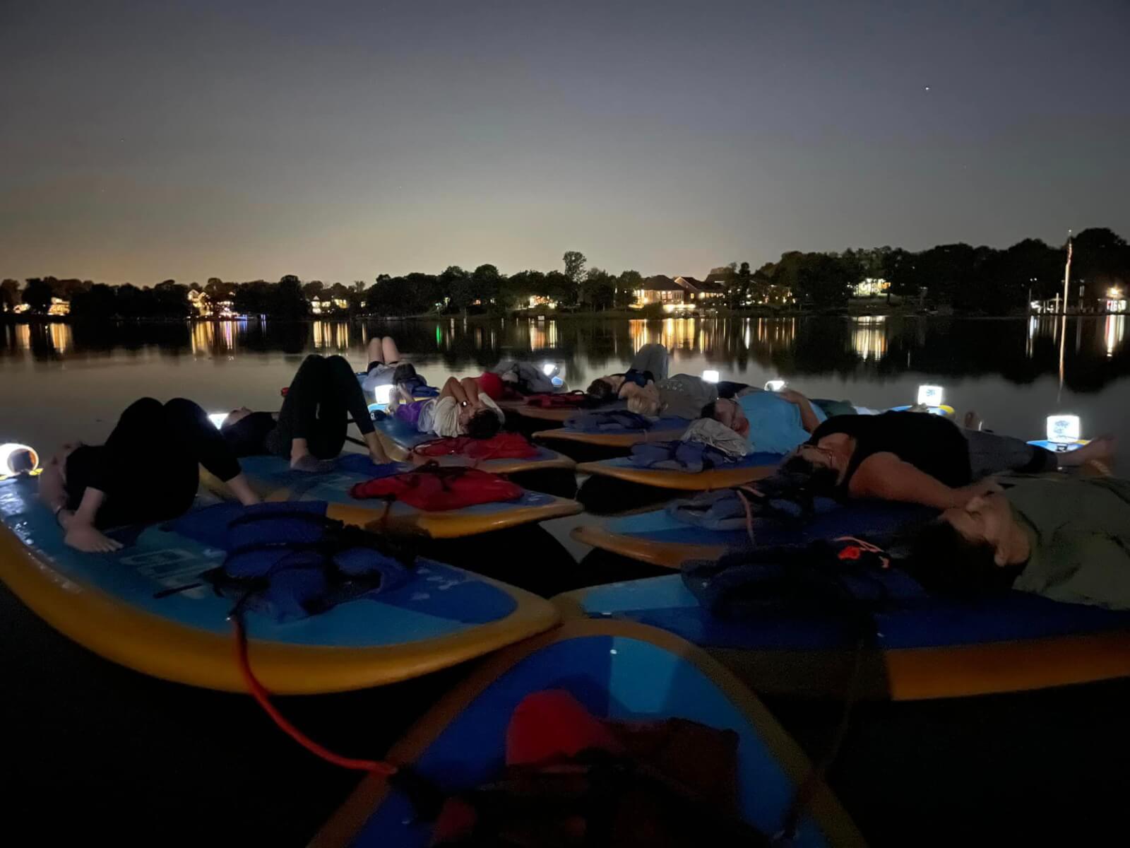 People relax on kayaks at night, floating on a calm lake under a dark sky with city lights in the background.