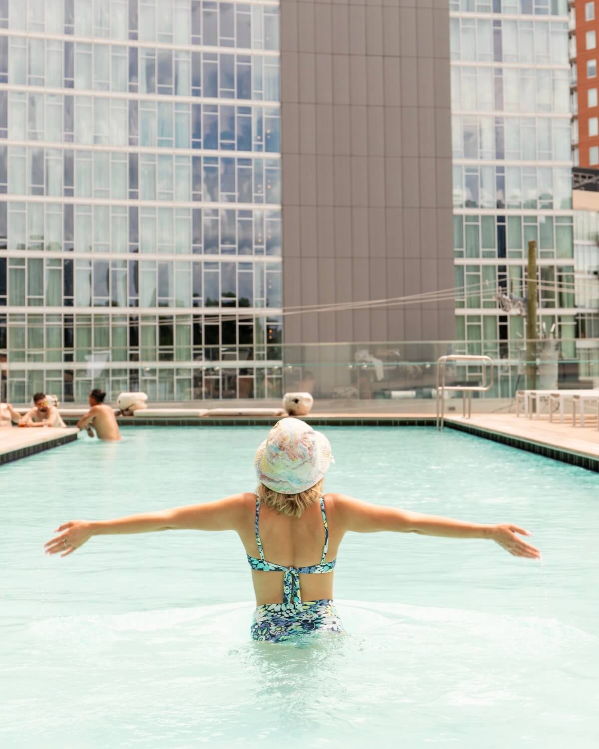 A person in a swimsuit and swim cap is standing with arms outstretched in a pool, surrounded by tall buildings. Other swimmers are visible in the background.