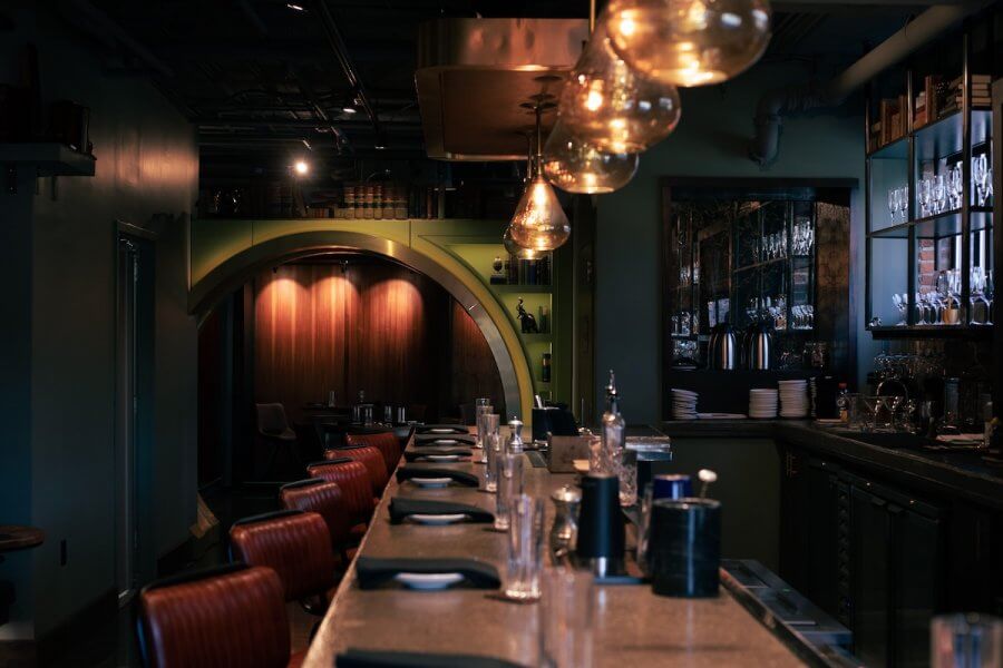 Interior of a dimly lit bar with leather stools, a wooden counter, elegant glassware on shelves, and large, glowing pendant lights.