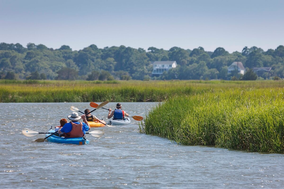 Four people kayaking on a serene river with lush greenery and a distant house, under a clear blue sky.