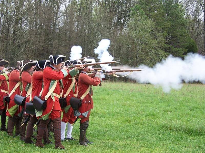 A group of historical reenactors dressed in red military uniforms firing muskets in a grassy field.