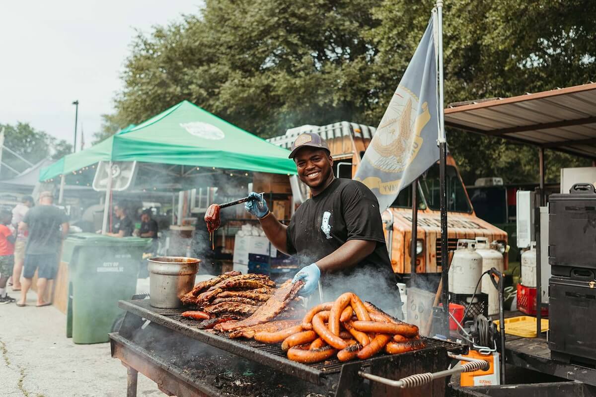 Man grilling sausages and ribs at an outdoor food stall, smiling at the camera, with people and tents in the background.