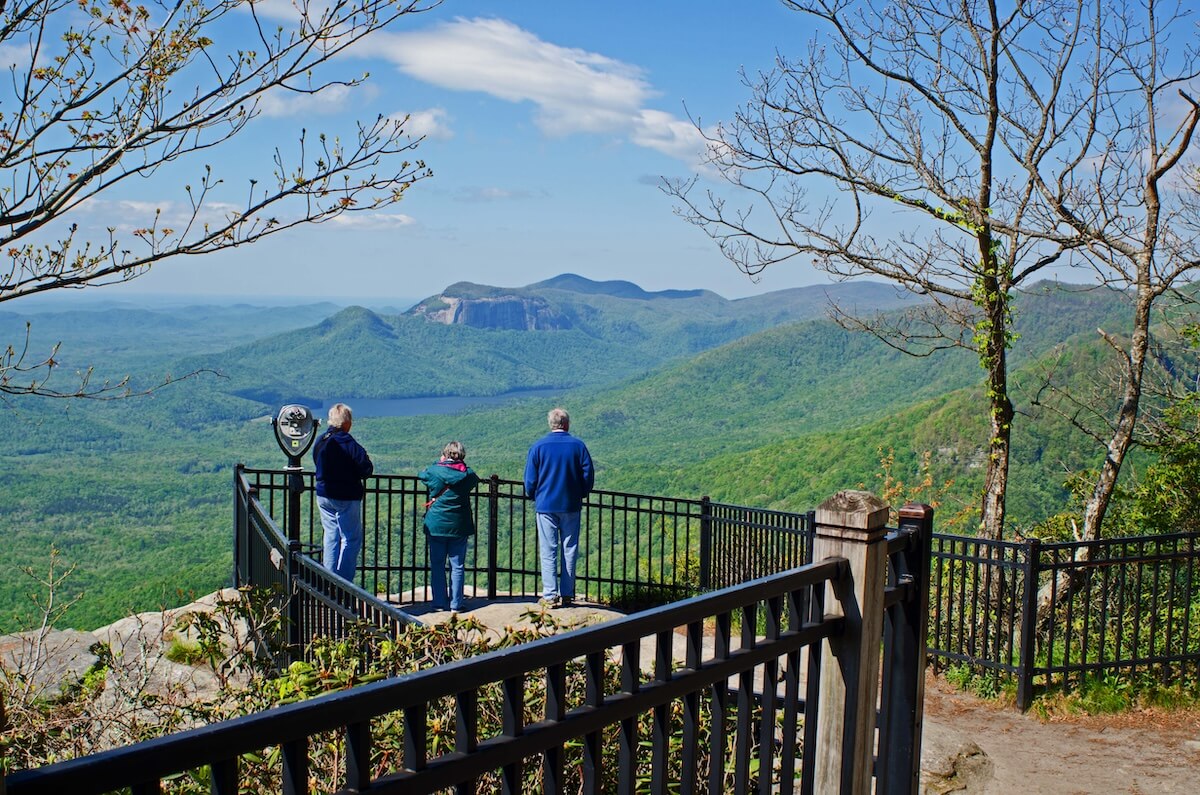Three people observing a scenic mountain view from an overlook with a metal railing, clear blue sky above and lush greenery below.