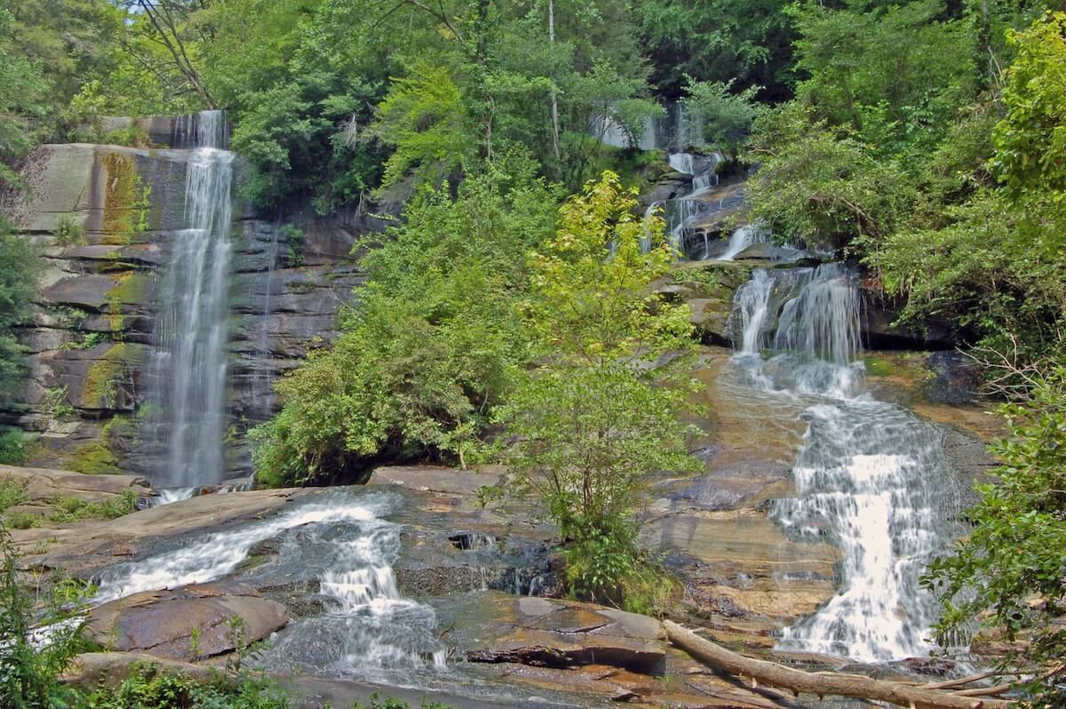 A multi-tiered waterfall cascades over smooth rock faces surrounded by lush, green forest under a clear sky.