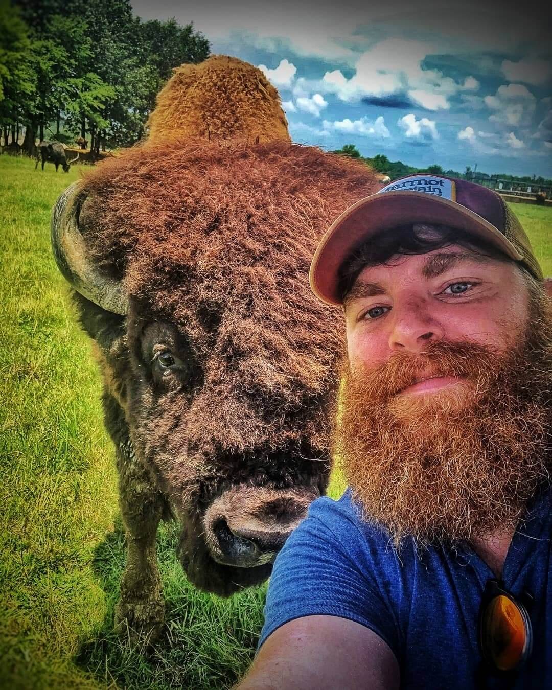 A man with a beard and a baseball cap takes a selfie with a buffalo in a grassy field during his visit to Tupelo, MS. Trees and a partly cloudy sky are visible in the background.