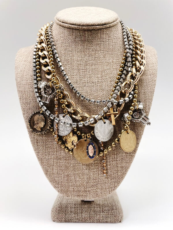 A mannequin bust displaying a layered necklace made of various chains and charms, including coins and crosses.