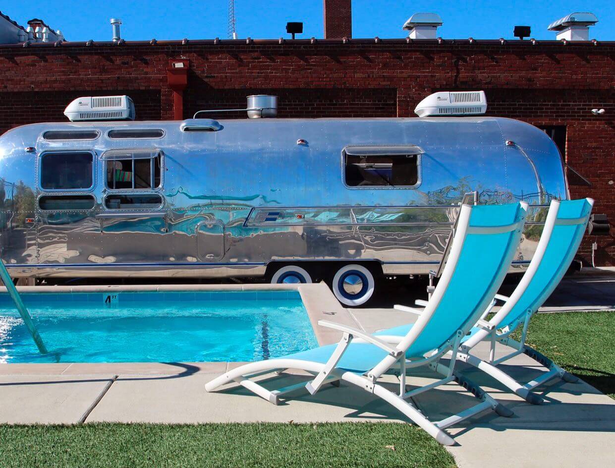 A shiny airstream trailer parked next to a swimming pool with two blue lounge chairs in the foreground.