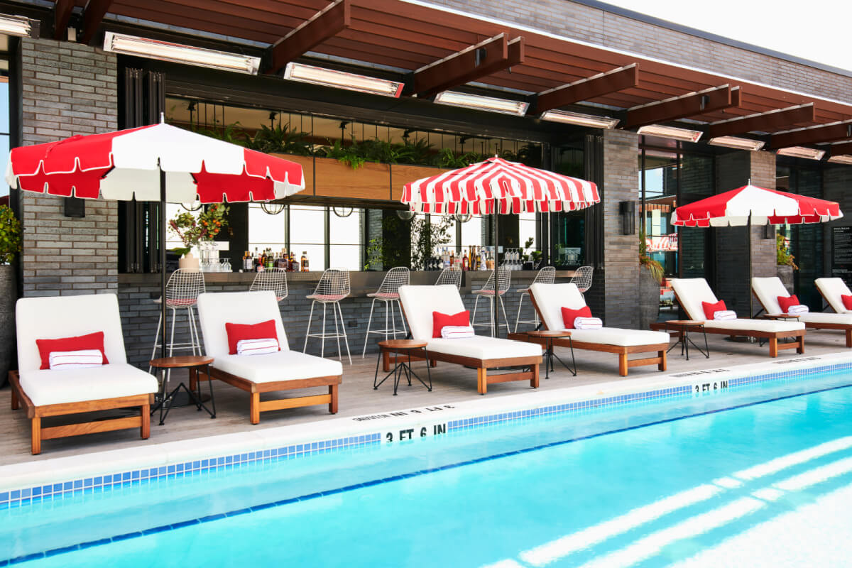 Outdoor pool area with white lounge chairs, red and white striped umbrellas, and a poolside bar, reflecting modern architecture.