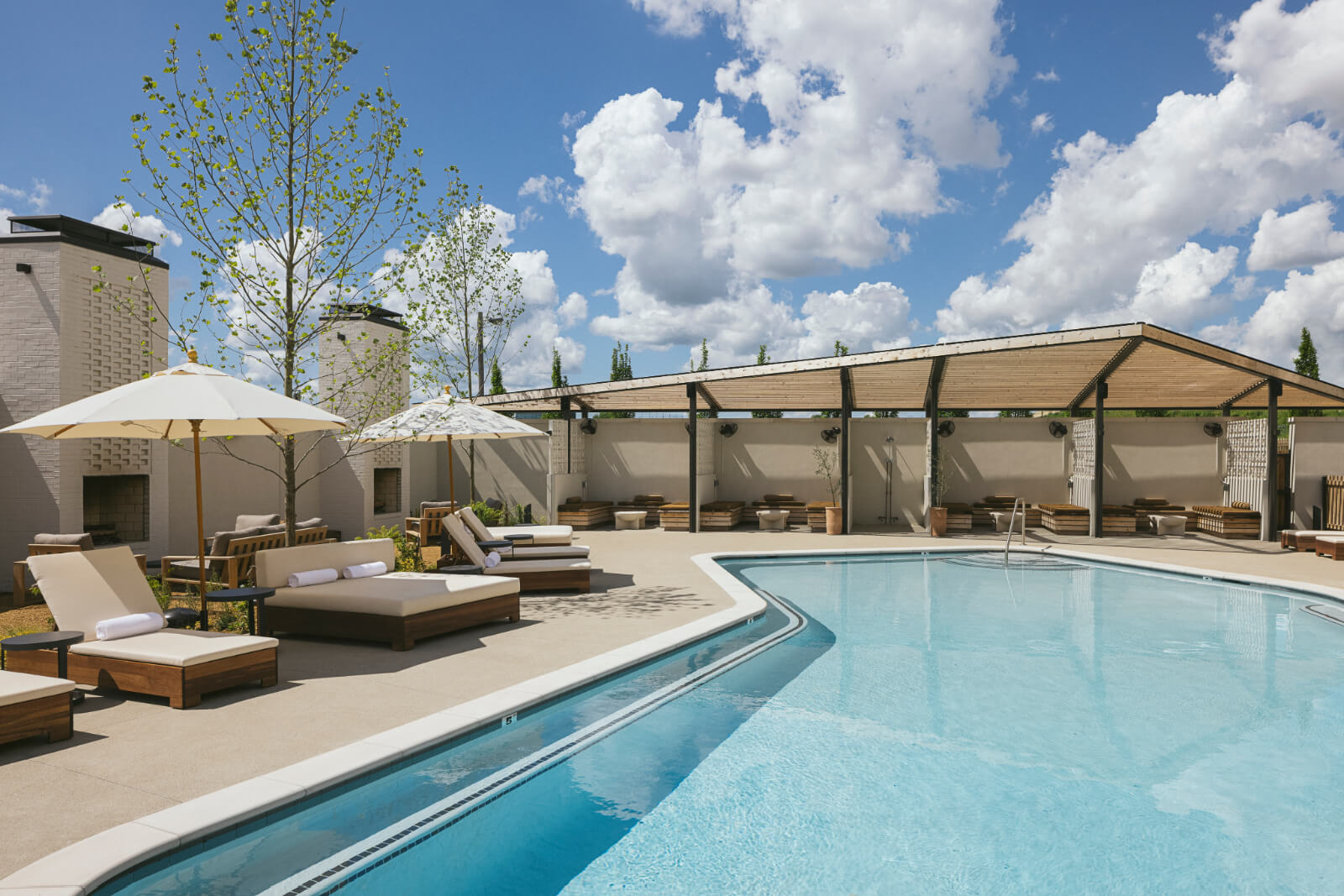 A clear blue outdoor swimming pool, typical of elegant hotel pools, is surrounded by lounge chairs and umbrellas, with a shaded seating area in the background under a partly cloudy sky.