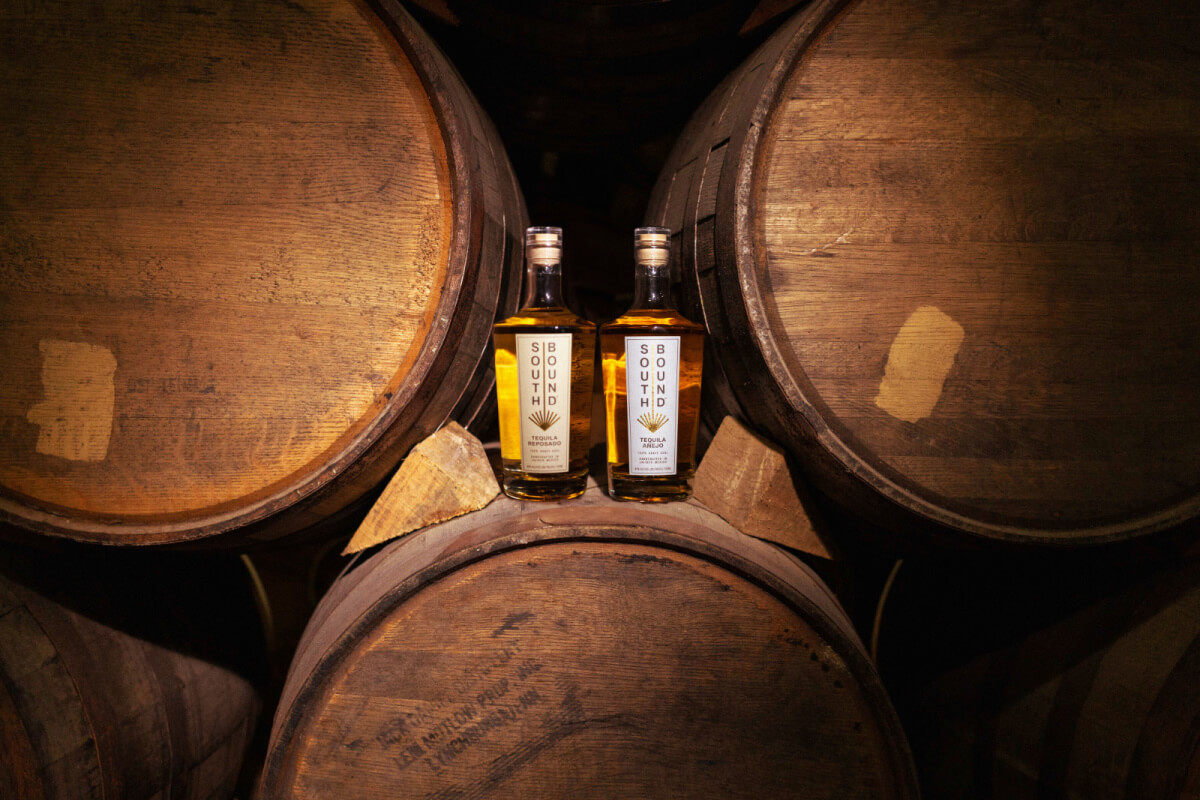 Two bottles of bourbon positioned between aged wooden barrels in a dimly lit storeroom.