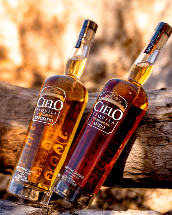 Two bottles of cielo tequila, reposado and anejo, resting on a wooden log with a blurred natural background.