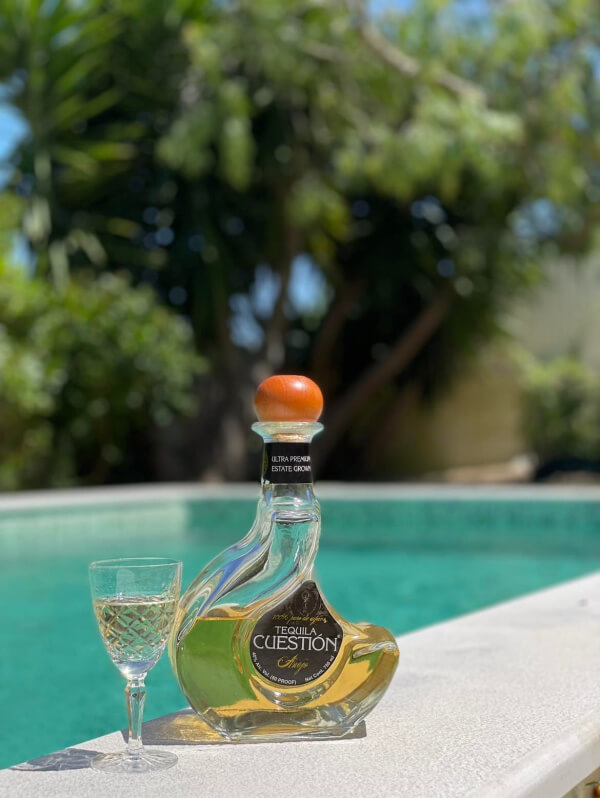 A bottle of cuestión tequila and a filled shot glass on a poolside ledge, with lush greenery in the background.