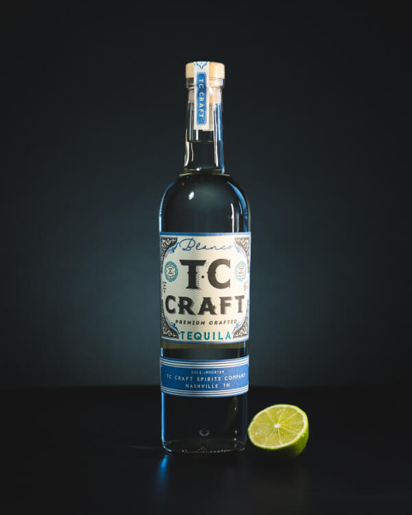 A bottle of tc craft tequila with a blue label, displayed against a black background, accompanied by a slice of lime.