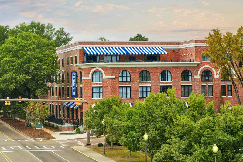 Red-brick building features arched windows and blue-striped awnings, surrounded by trees. The building proudly displays a sign reading "GRADUATE" on one side and is located near a street intersection.