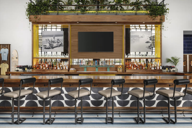 A modern bar with a wooden counter, six high chairs, a large central TV, various bottles on display, and framed black-and-white photos. Green plants adorn the top shelf. 