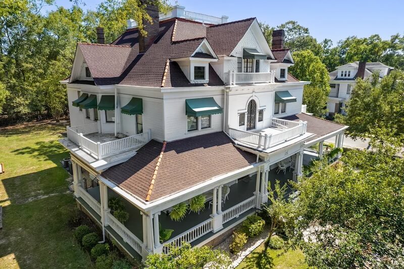 A large, two-story white house with green window awnings, multiple gables, and a wraparound porch, surrounded by Mississippi's lush trees and greenery.