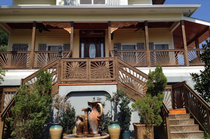A charming two-story house in Mississippi, boasting an elaborate wooden front porch and staircase. The entrance features a carved wood design, and a decorative fountain with jugs graces the front yard. It's the perfect boutique hotel experience nestled in Southern elegance.