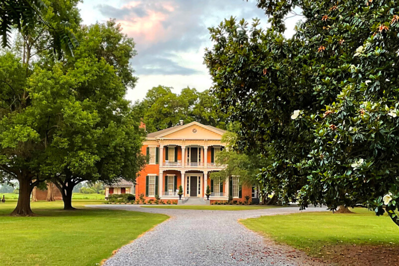 A large two-story house with a columned porch sits at the end of a gravel driveway, surrounded by trees and greenery under a partly cloudy Mississippi sky, reminiscent of charming boutique hotels.