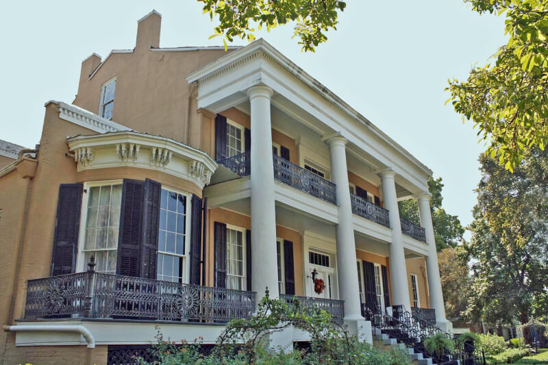 A top historical two-story house in Mississippi featuring large white columns, a wrap-around balcony with wrought-iron railings, and multiple windows, surrounded by trees and landscaping.