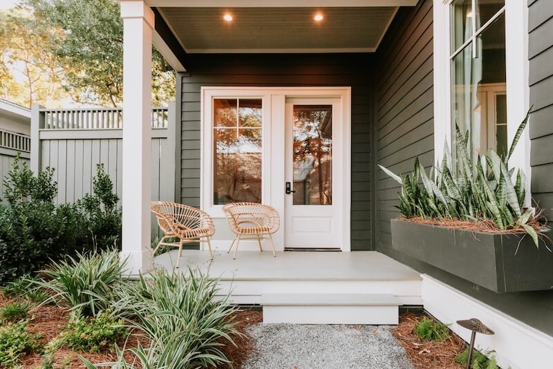 A front porch with two rattan chairs, a white door with glass panels, a large window, a planter box with succulents, and green siding. The porch is surrounded by greenery.