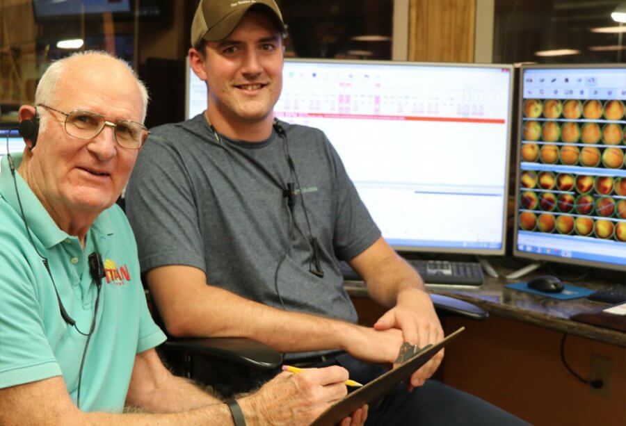 Two men, one older and one younger, sit at a desk with computer monitors displaying data and images of Titan Farms peaches in the background.