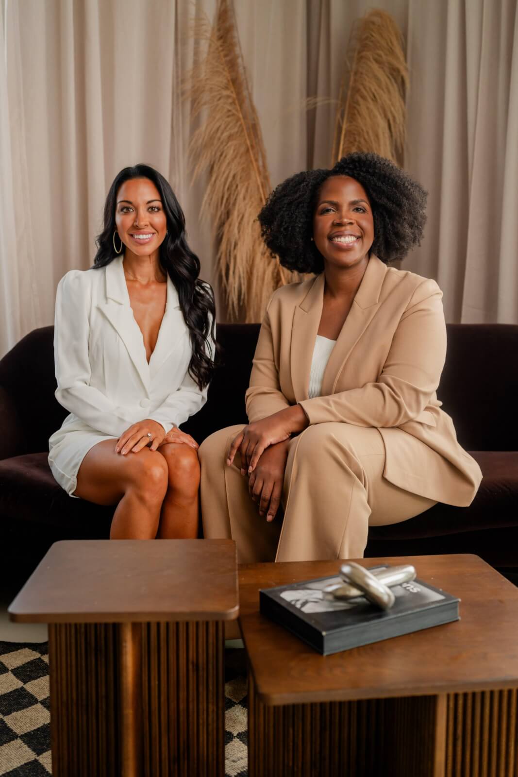 Two women seated on a sofa, one in a white outfit and the other in a beige suit, smile towards the camera. The elegant setting, reminiscent of the top med spas in Nashville, features simple decor and pampas grass softly adorning the background.