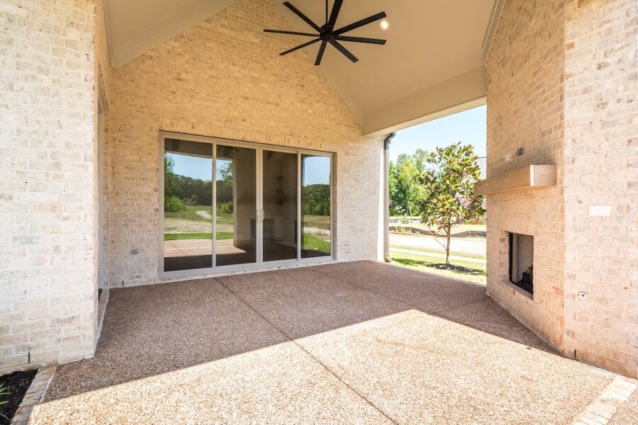 A covered patio at 140 Magnolia Ridge features a large ceiling fan, sliding glass doors, and a built-in fireplace. The floor is made of speckled concrete, with light-colored brick walls. Trees and a lawn are visible outside, creating a serene Rossville, Tennessee backdrop.