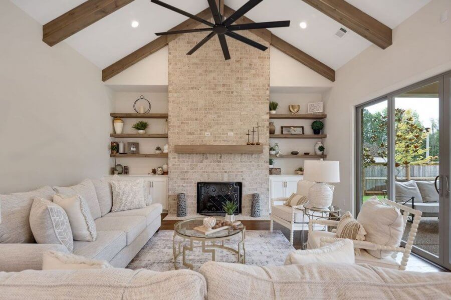 Modern living room at 140 Magnolia Ridge Drive, Rossville Tennessee, with vaulted ceiling, exposed wood beams, large ceiling fan, brick fireplace, built-in shelves, neutral furniture, and glass doors leading to a patio.