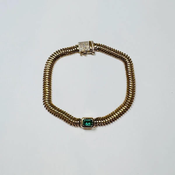 A gold bracelet with a ridged design, from our luxe loves collection, featuring a rectangular green gemstone set in the center and a clasp adorned with small clear stones.