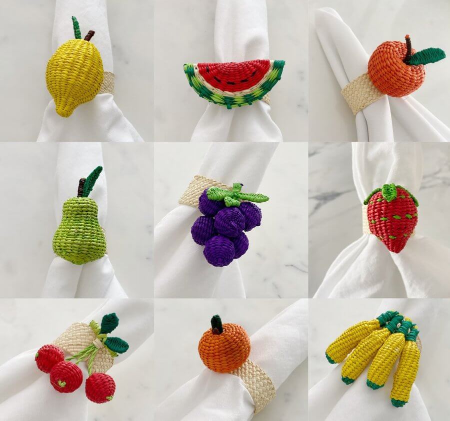 A 3x3 grid of woven rattan fruit-themed napkin rings, each showcasing a different fruit: lemon, watermelon, apple, pear, grapes, strawberry, cherries, orange, and bananas. All are attached to white napkins.