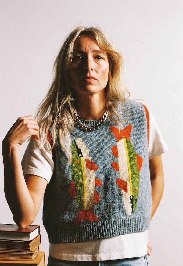 A woman with long blonde hair wears a knitted vest featuring two colorful fish designs, over a white t-shirt—serving luxe loves vibes as she poses beside a stack of books against a plain background.