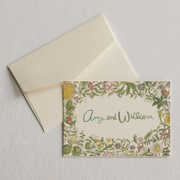 A cream-colored envelope and a floral-bordered card with "Amy and William" written in green cursive are shown on a light fabric background, perfectly capturing the essence of luxe loves.