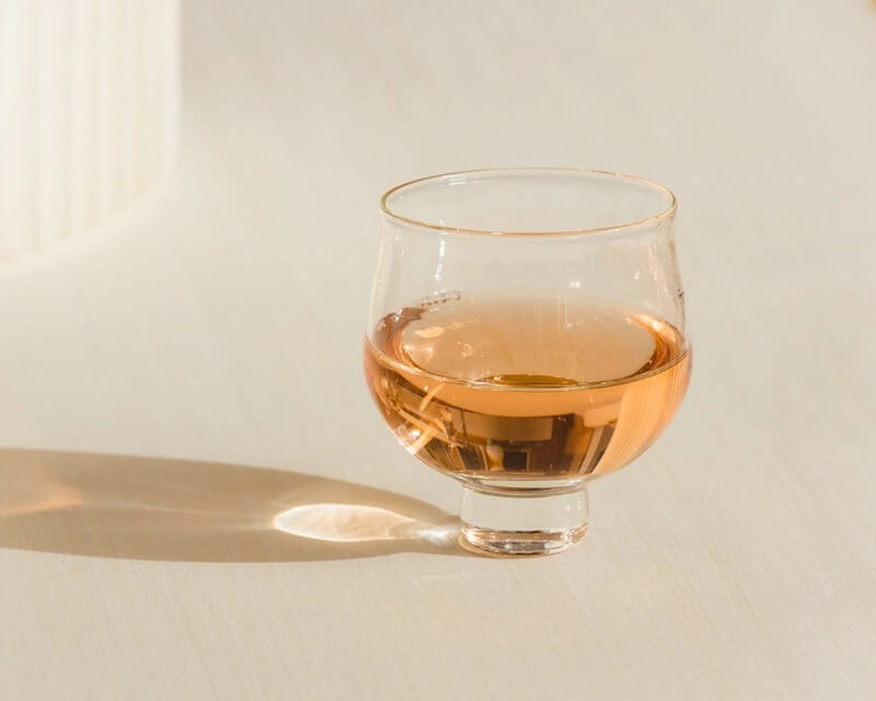 A clear glass filled with a light amber liquid sits on a light surface, casting a soft shadow to the left—perfect for moments when luxe loves simplicity.