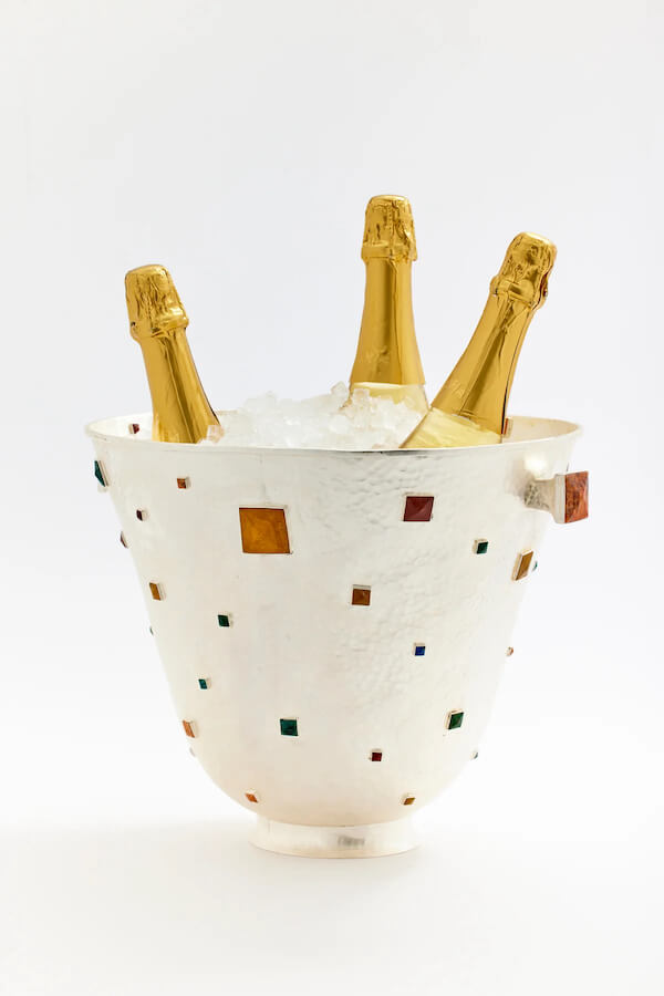 A white ice bucket with colored square accents holds three bottles of champagne, each wrapped in gold foil, surrounded by ice—perfect for luxe loves celebrating in style.