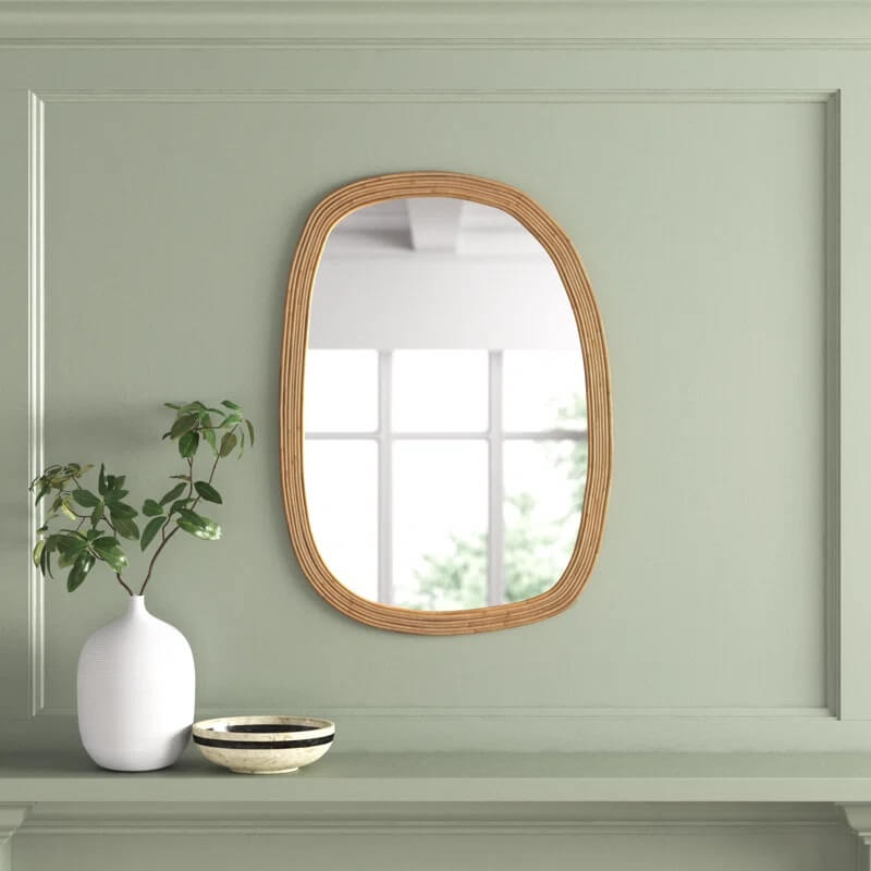 A wall-mounted mirror with a light wooden rattan frame hangs above a mantel decorated with a white vase containing greenery and a small ceramic bowl.