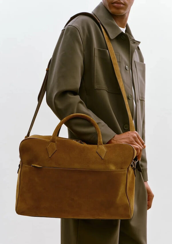A person wearing an olive-green outfit holds a brown suede leather duffel bag—one of this season's luxe loves—with both handles and a shoulder strap.
