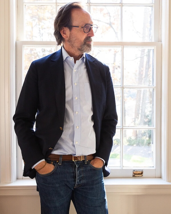 A man with glasses and a beard stands in front of a window, wearing a dark blazer, light blue shirt, and jeans with hands in his pockets—embodying the effortless style luxe loves.