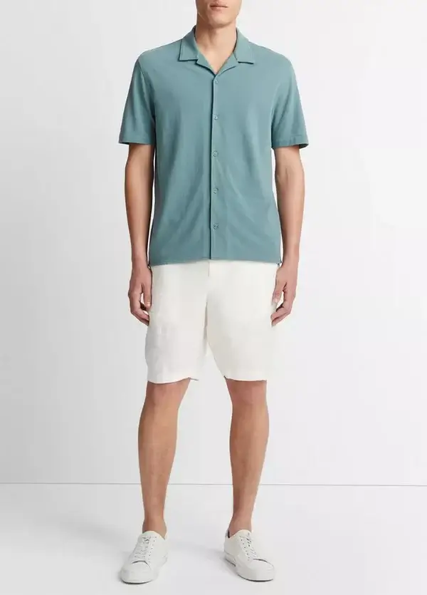 A person is wearing a teal short-sleeve button-up shirt, cream shorts, and white sneakers, standing against a plain background, embodying effortless style that could spark inspiration for expensive gift ideas.