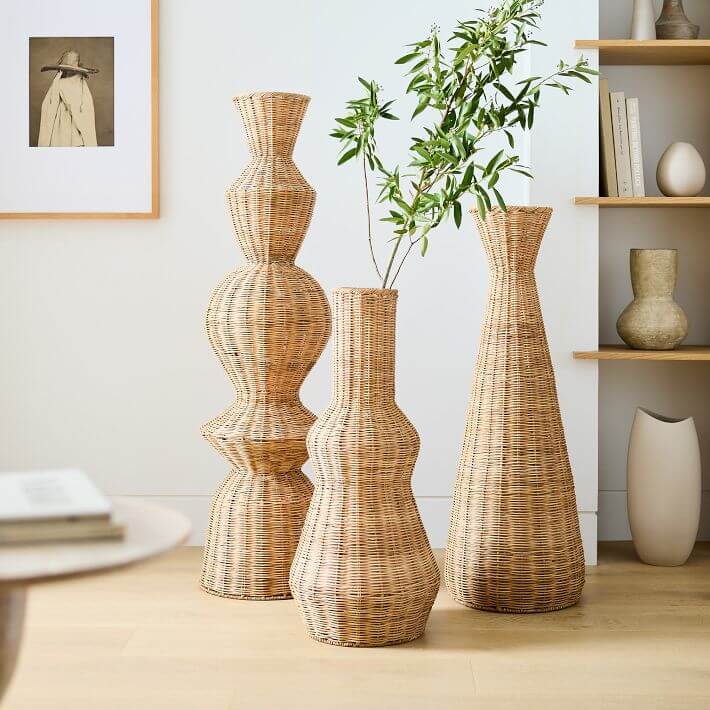 Three large wicker vases of different shapes, including one with a small leafy branch, are placed on a wooden floor adjacent to a bookshelf and a framed picture. The rattan design adds an elegant touch to the cozy arrangement.