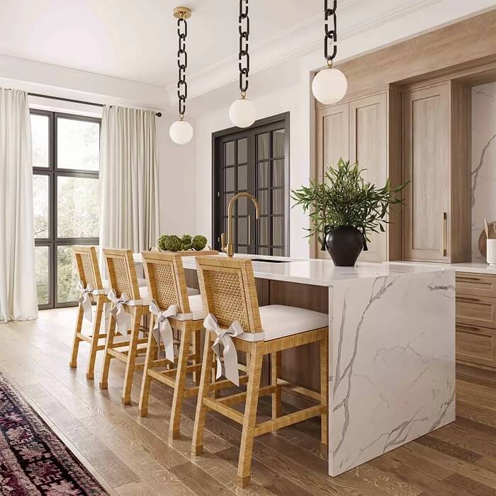 Modern kitchen featuring a marble island with rattan barstools, pendant lights, white cabinetry, large windows with curtains, and a potted plant centerpiece.