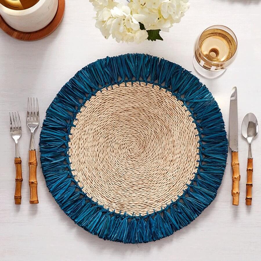 A round rattan woven placemat with blue fringe, set on a table with two forks on the left, a knife and spoon on the right, a glass of white wine, and a white floral arrangement in the background.