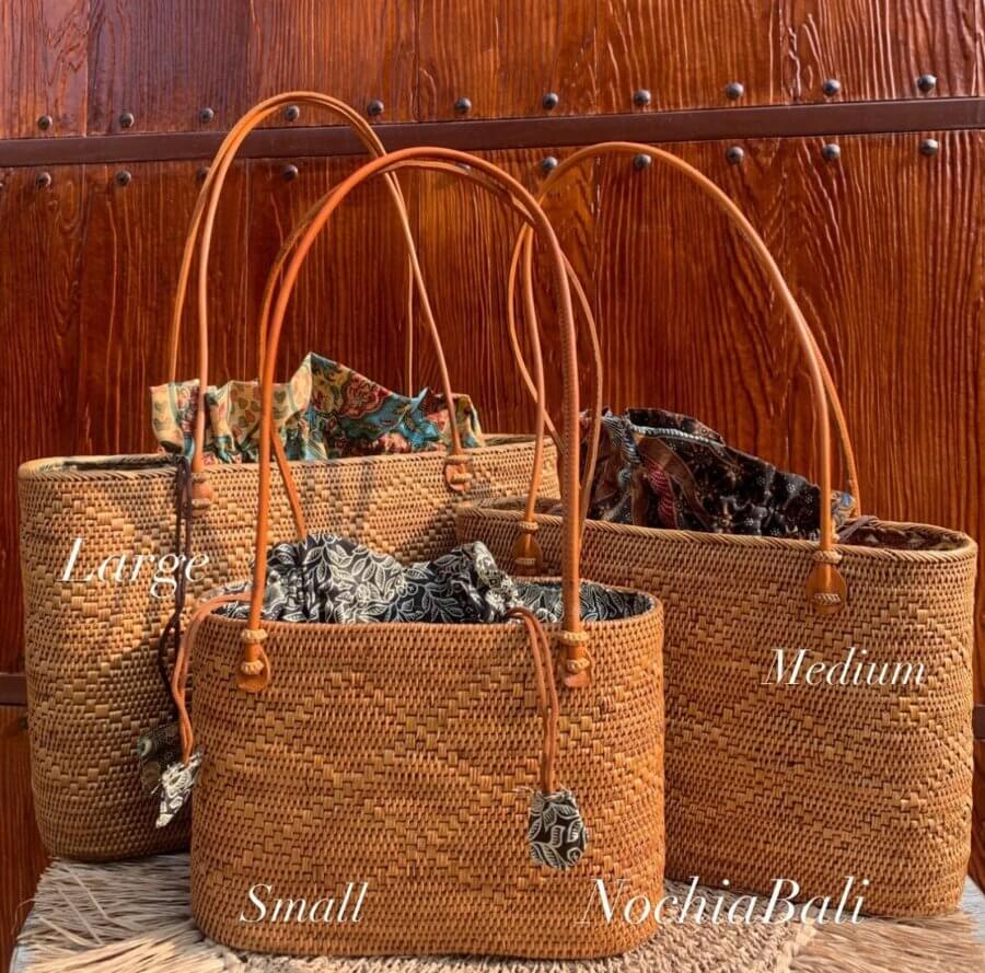Three woven straw baskets with leather handles, labeled by size as large, medium, and small, are displayed in front of a wooden background. Fabric is peeking out from each rattan basket.
