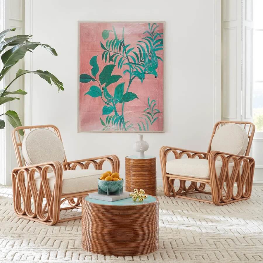 A room features two cozy rattan chairs with cushions, a small rattan side table, and a matching coffee table adorned with a plant and bowl. A pink and green leafy artwork hangs on the wall.