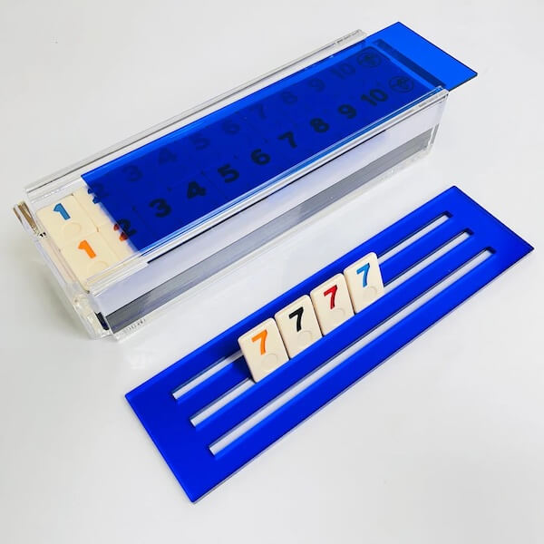 A rectangular game tray holds tiles numbered 1 to 13, with a blue cover. Four tiles—three orange 7s and one blue 7—stand upright on a separate blue rack, perfect for those who appreciate the little luxe loves in classic games.