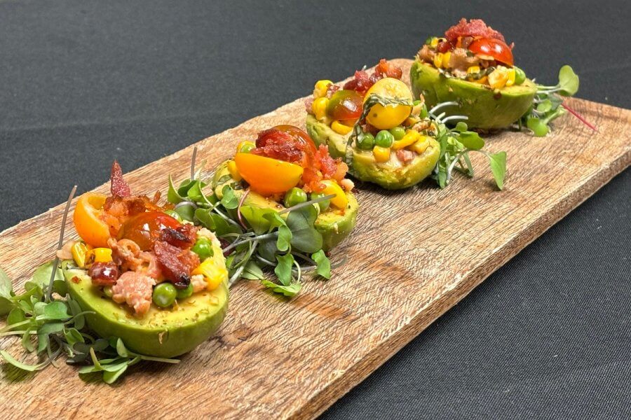 Four halved avocados, or avocado cups, are filled with a colorful mix of peas, corn, tomatoes, and bacon bits. They are beautifully arranged on a wooden serving board and garnished with fresh microgreens for an easy summer vegetable salad recipe.