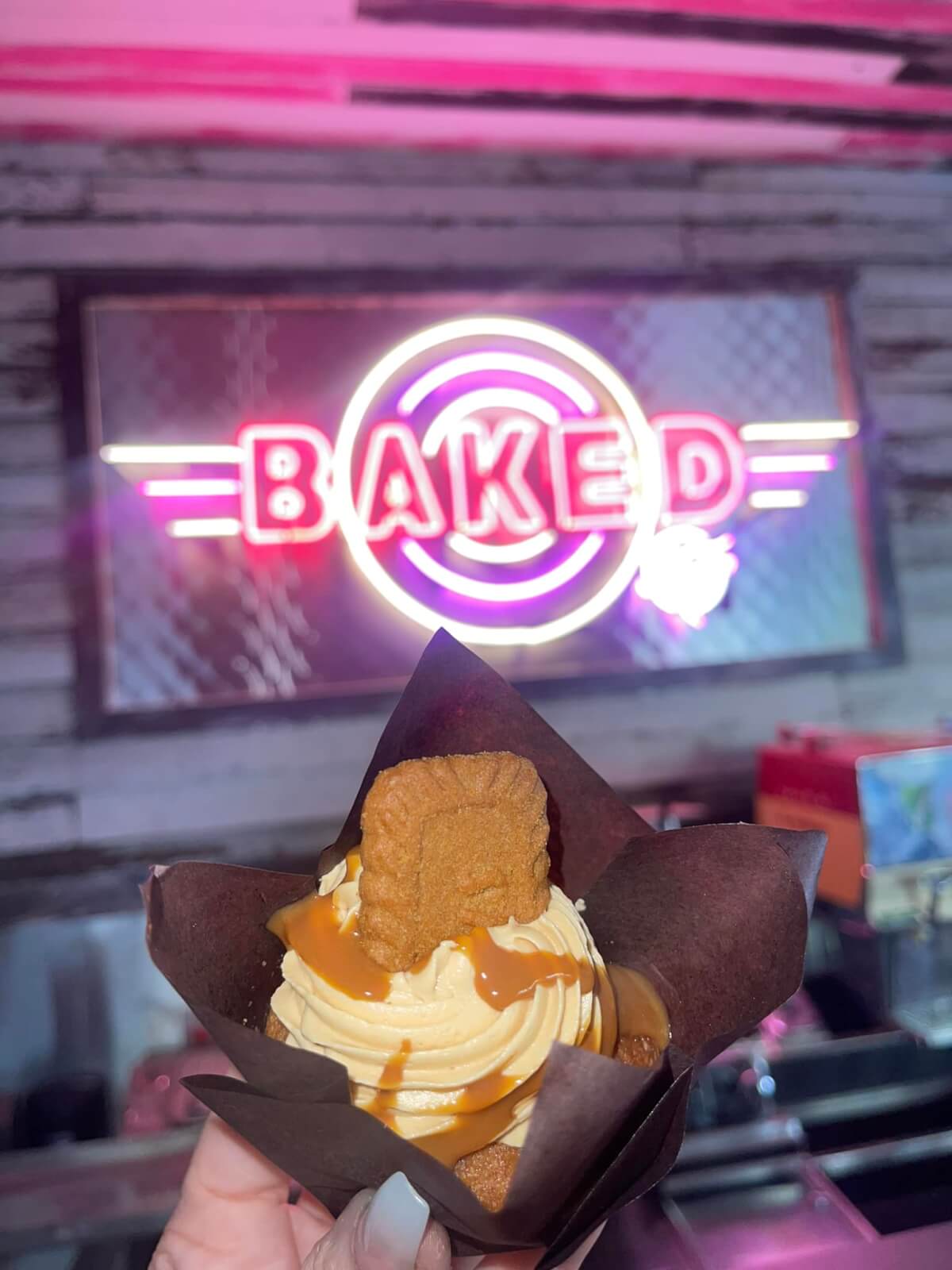 A hand holding a caramel cupcake with cookie topping in front of a neon 