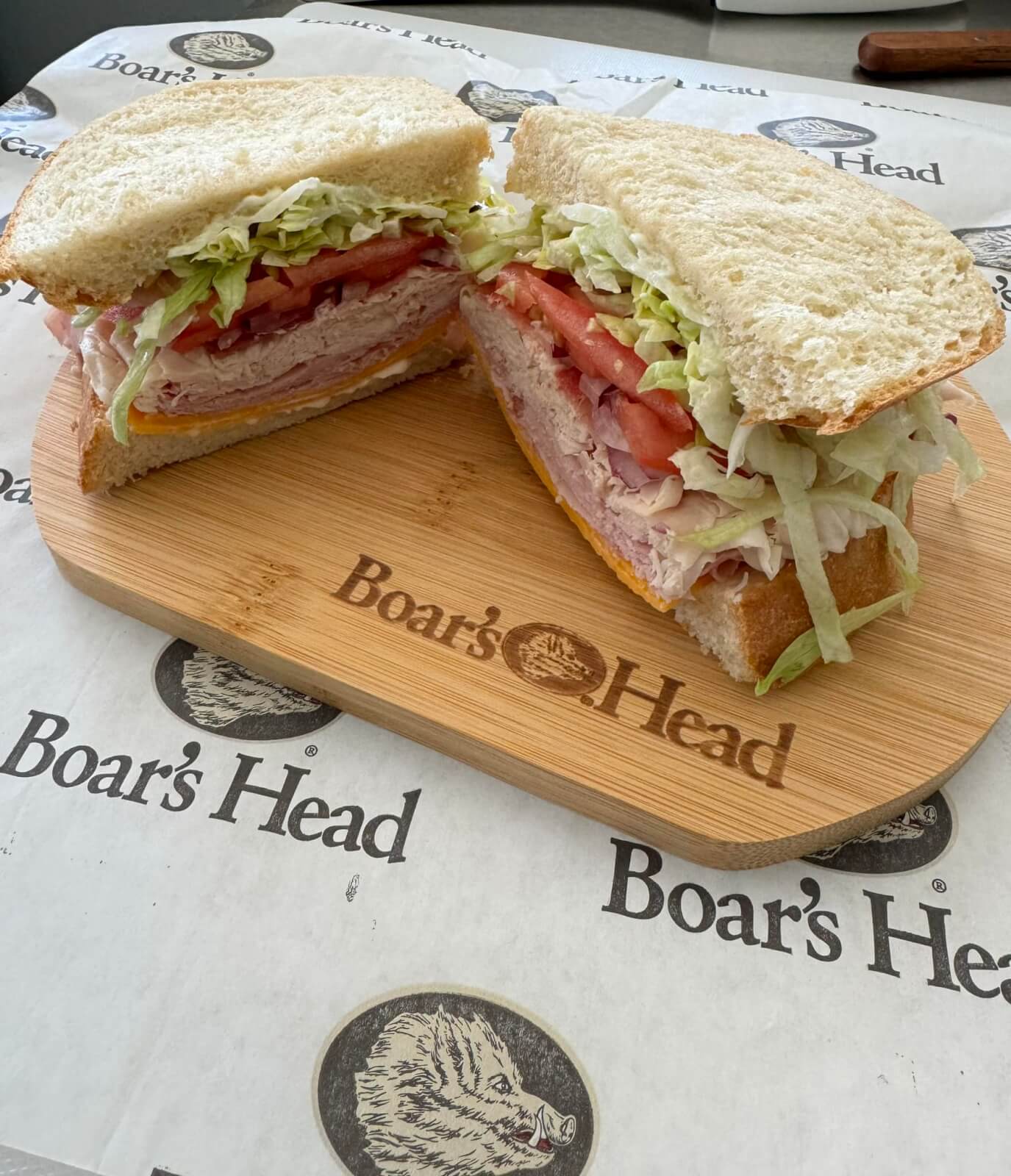 A sandwich cut in half, filled with sliced meat, tomato, lettuce, and cheese, placed on a wooden cutting board with 