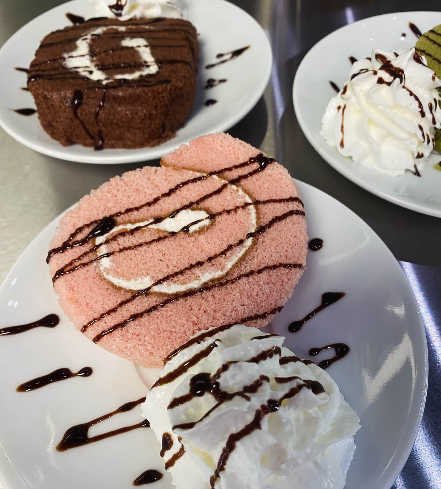 A plate with a pink Swiss roll, whipped cream, and chocolate drizzle in the foreground. Two similar desserts are in the background, one chocolate roll and the other undecorated with whipped cream.